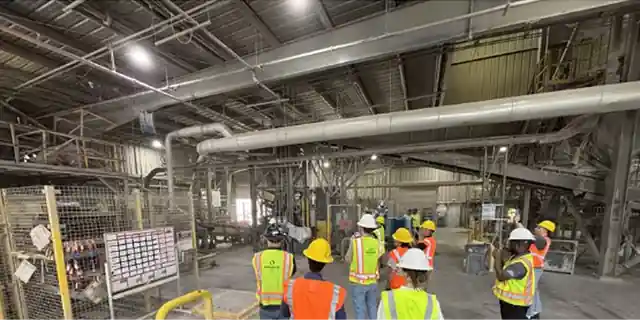 A group in hard hats and hi-vis vests gathered on the Oldcastle APG plant floor beside a guarded production line