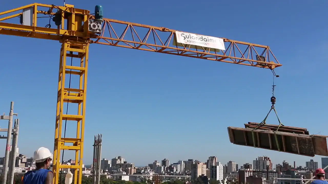 Tower crane lifting a slung load of boards above a rooftop as a worker in a hard hat looks on